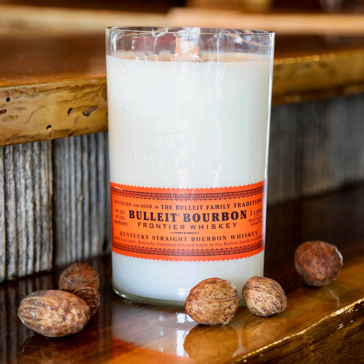 A Recycled Kentucky Bourbon Whiskey Candle in a clear glass with an orange label sits on a wooden surface next to several whole nutmegs.