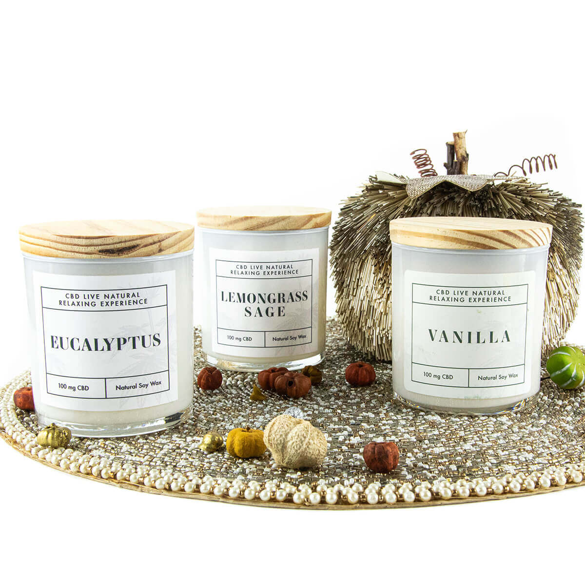 Three glass candles—Eucalyptus, LEMONGRASS SAGE Relaxing Aromatherapy Candle 11oz, and Vanilla—sit on a decorative tray with small artificial pumpkins and a textured ornament in the background for a calming candle display.
