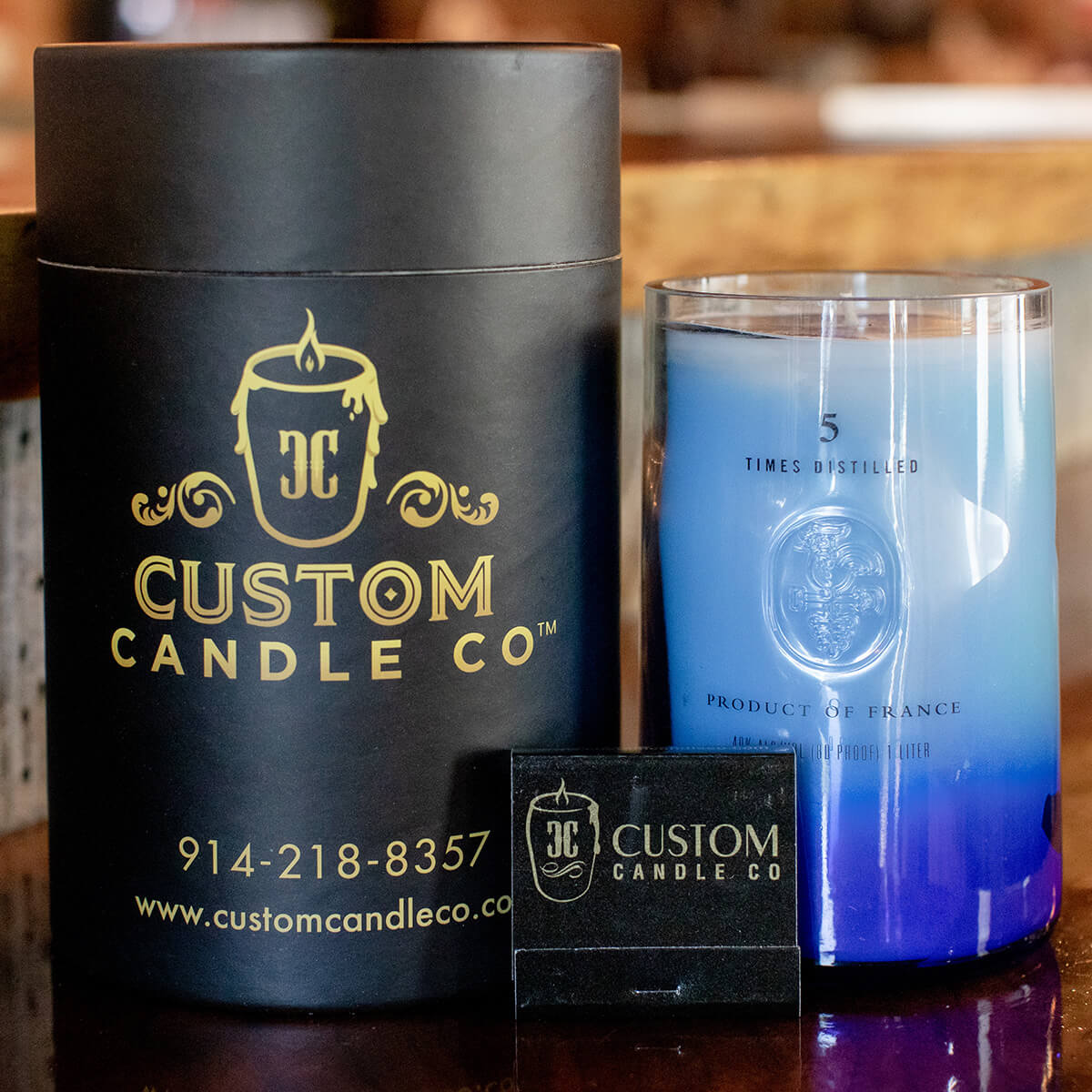 A Recycled Vodka Candle – Multi-color in a glass jar sits next to a black cylindrical Custom Candle Co box and a black business card on a wooden surface.