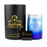A Recycled Vodka Candle – Multi-color in a glass jar is displayed beside a black cylindrical "Custom Candle Co" box and a small black matchbox.