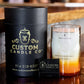A Recycled Vodka Candle – Multi-color sits in a glass holder beside its black cylindrical packaging and a small matchbox, all displayed on a wooden surface.