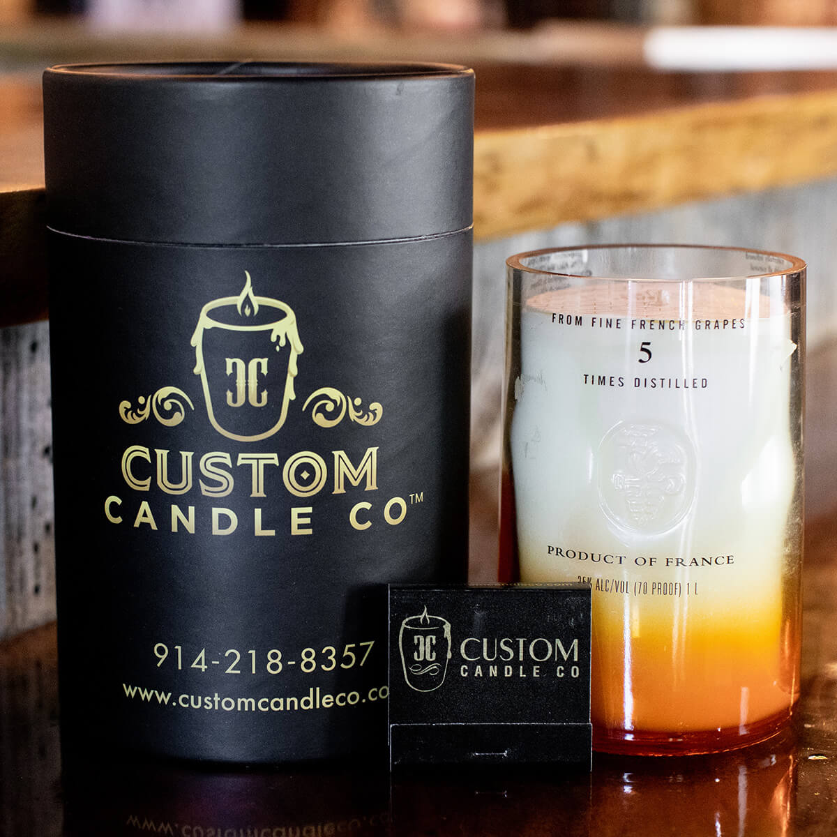 A Recycled Vodka Candle – Multi-color sits in a glass holder beside its black cylindrical packaging and a small matchbox, all displayed on a wooden surface.