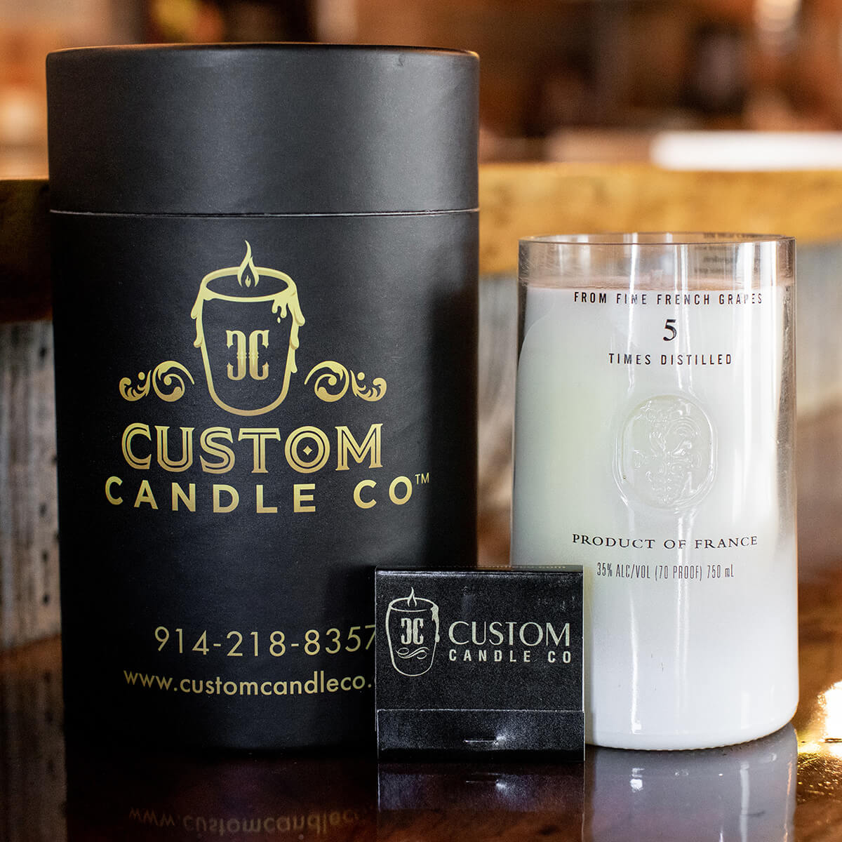 A Recycled Vodka Candle – Multi-color is displayed in a clear glass jar next to a black cylindrical box and a small black matchbox on a wooden surface.