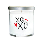 The XOXO Colored Quote Candle is hand poured with white essential oils in a clear tumbler glass jar, topped with a silver lid and featuring "xo XO" and three red hearts on the front.