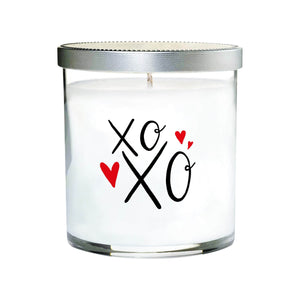 The XOXO Colored Quote Candle is hand poured with white essential oils in a clear tumbler glass jar, topped with a silver lid and featuring "xo XO" and three red hearts on the front.