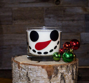 A Snowman 2 Wick Candle sits on a cut birch log beside small red and green Christmas ornaments, against a wooden background—perfect for adding festive holiday scents to your Merry Christmas display.
