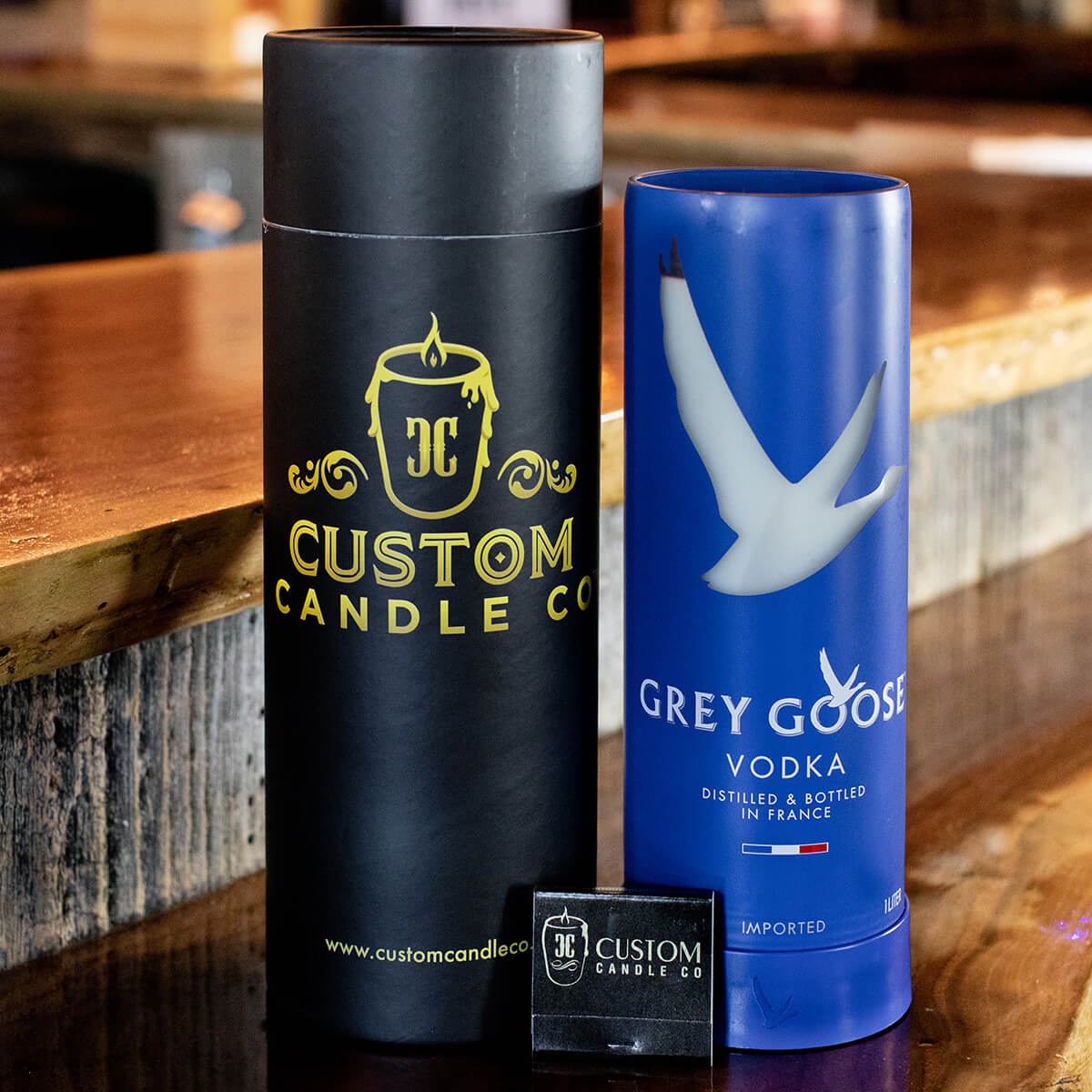 A Recycled French Vodka Candle - Blue sits on a wooden surface next to a Grey Goose bottle, with a business card in front.