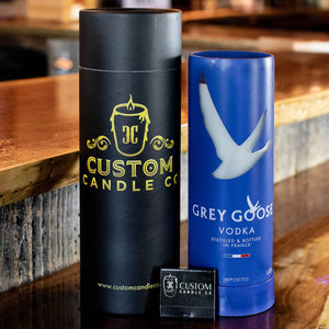 A Recycled French Vodka Candle - Blue sits on a wooden surface next to a Grey Goose bottle, with a business card in front.