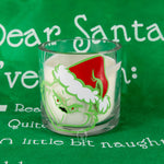 The Grinch Holiday Candle is a clear glass Christmas candle featuring the Grinch in a Santa hat, placed before a green cloth with white text about Santa—a festive gift idea for any Grinch fan.