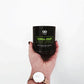 A hand holds the CHILL-OUT Aromatherapy Candle 11oz, a black green tea lemongrass CBD candle made from all-natural soy wax. The person wears a watch with a tan strap and white face.