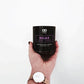 A hand holds the RELAX Aromatherapy Candle 11oz, a black CBD-infused candle with lavender and chamomile, against a plain white background. The person is wearing a watch with a brown strap.