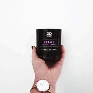 A hand holds the RELAX Aromatherapy Candle 11oz, a black CBD-infused candle with lavender and chamomile, against a plain white background. The person is wearing a watch with a brown strap.