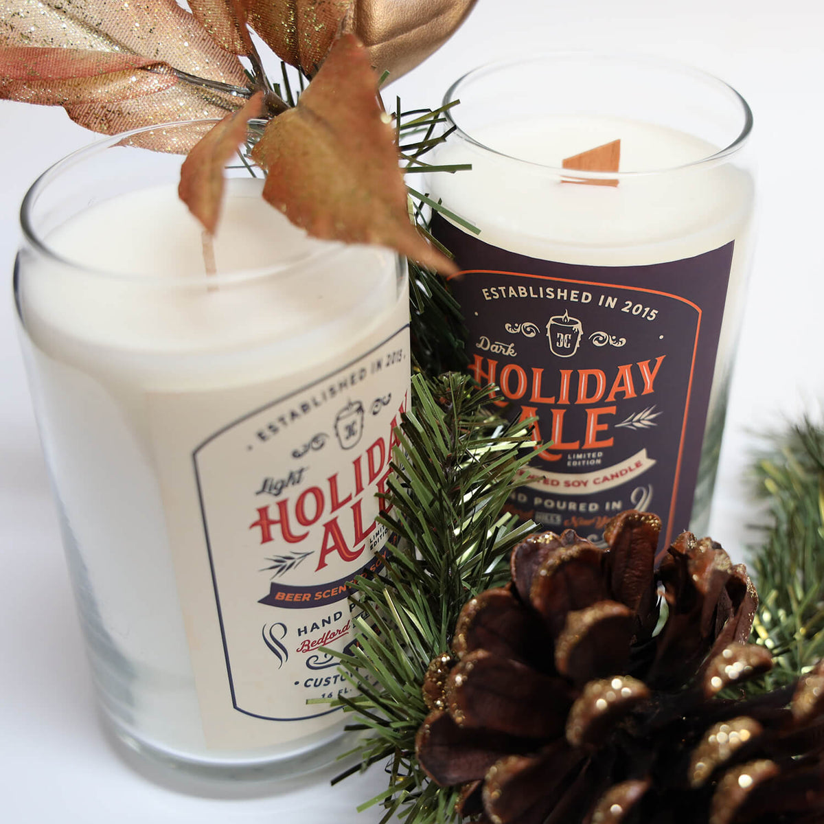 Two hand-poured Dark Holiday Ale Candles sit among pine branches, a pine cone, and decorative leaves on a white background.