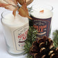 Two hand-poured Light Holiday Ale Candles with festive labels sit by pine branches and a pine cone, one adorned with a gold decorative leaf. Made from natural soy, these candles bring a holiday touch to any setting.