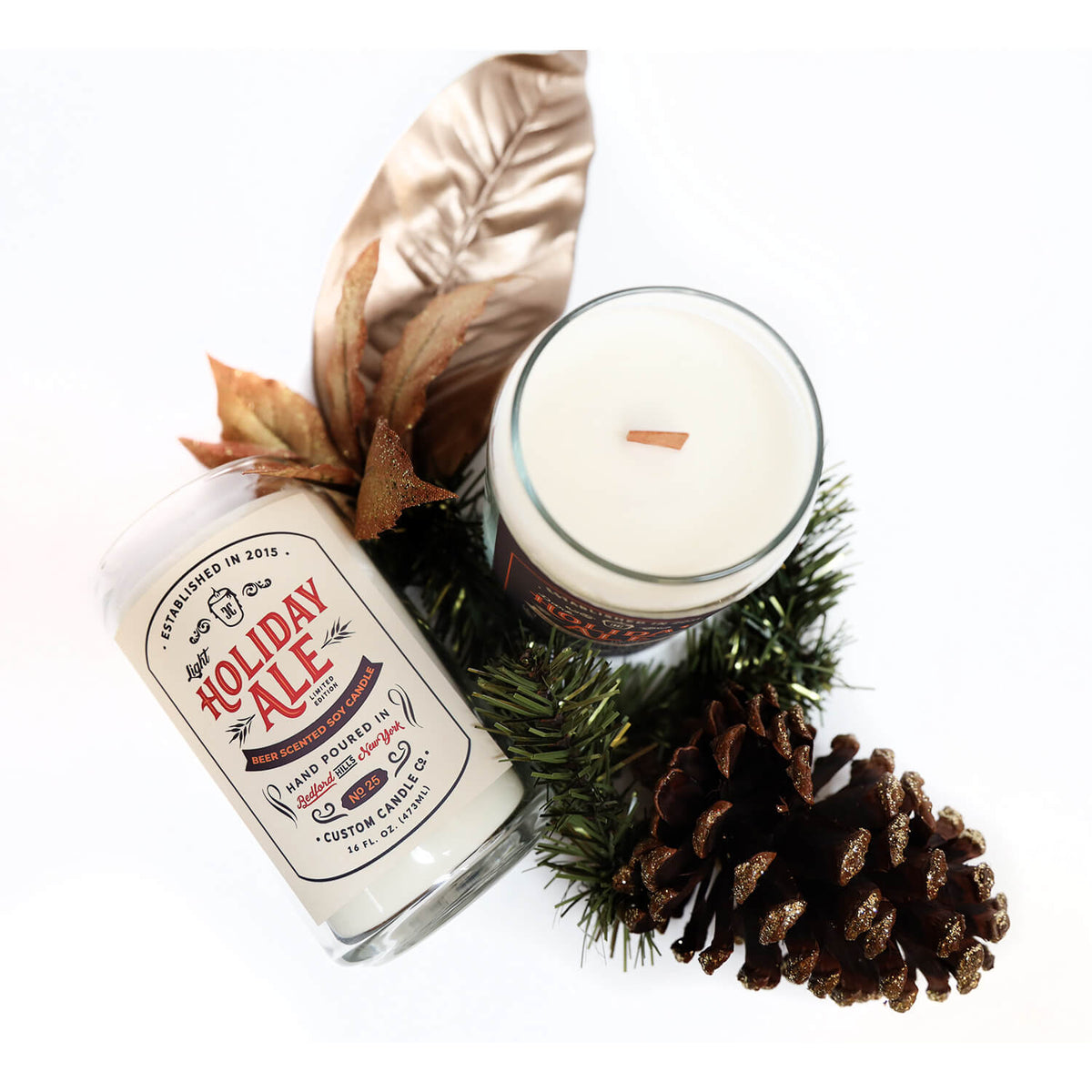 The Dark Holiday Ale Candle, a hand-poured soy candle with a wooden wick, is displayed beside pinecones, green pine branches, and a metallic leaf ornament for a cozy seasonal touch.