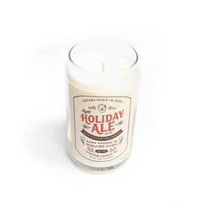The Light Holiday Ale Candle is a hand-poured natural soy candle in a clear glass with “Holiday Ale” label and decorative text, displayed on a white background.