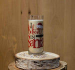 The "I Believe in Santa Claus Candle," made from a recycled liquor bottle, rests on a wooden slice base with clear decorative stones around it, all set against a wooden backdrop.
