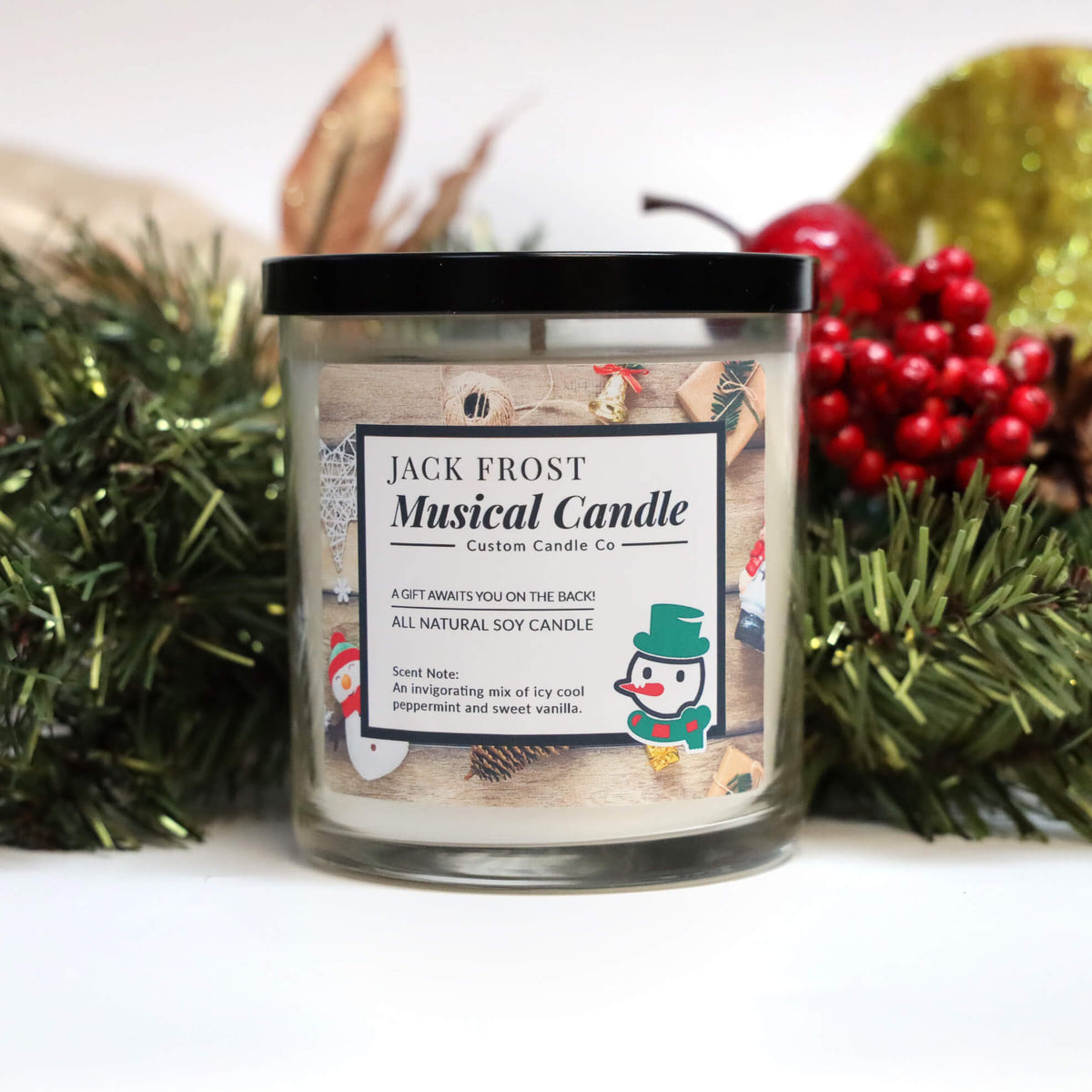 The Musical Holiday Candle & Diffuser | Jack Frost comes in a glass jar with a black lid, surrounded by pine branches, red berries, and a gold ornament.