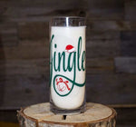 A clear glass labeled "Jingle" in green script with a red Santa hat on the "J," filled with milk on a wooden surface—perfect alongside your Jingle Ball Candle for festive holiday vibes.