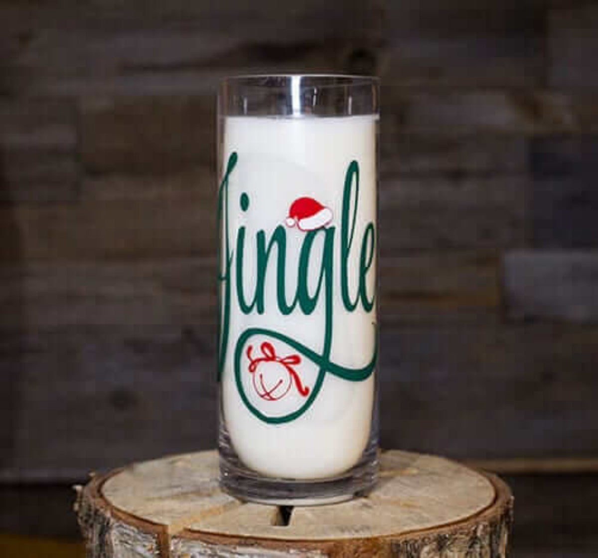 A clear glass labeled "Jingle" in green script with a red Santa hat on the "J," filled with milk on a wooden surface—perfect alongside your Jingle Ball Candle for festive holiday vibes.
