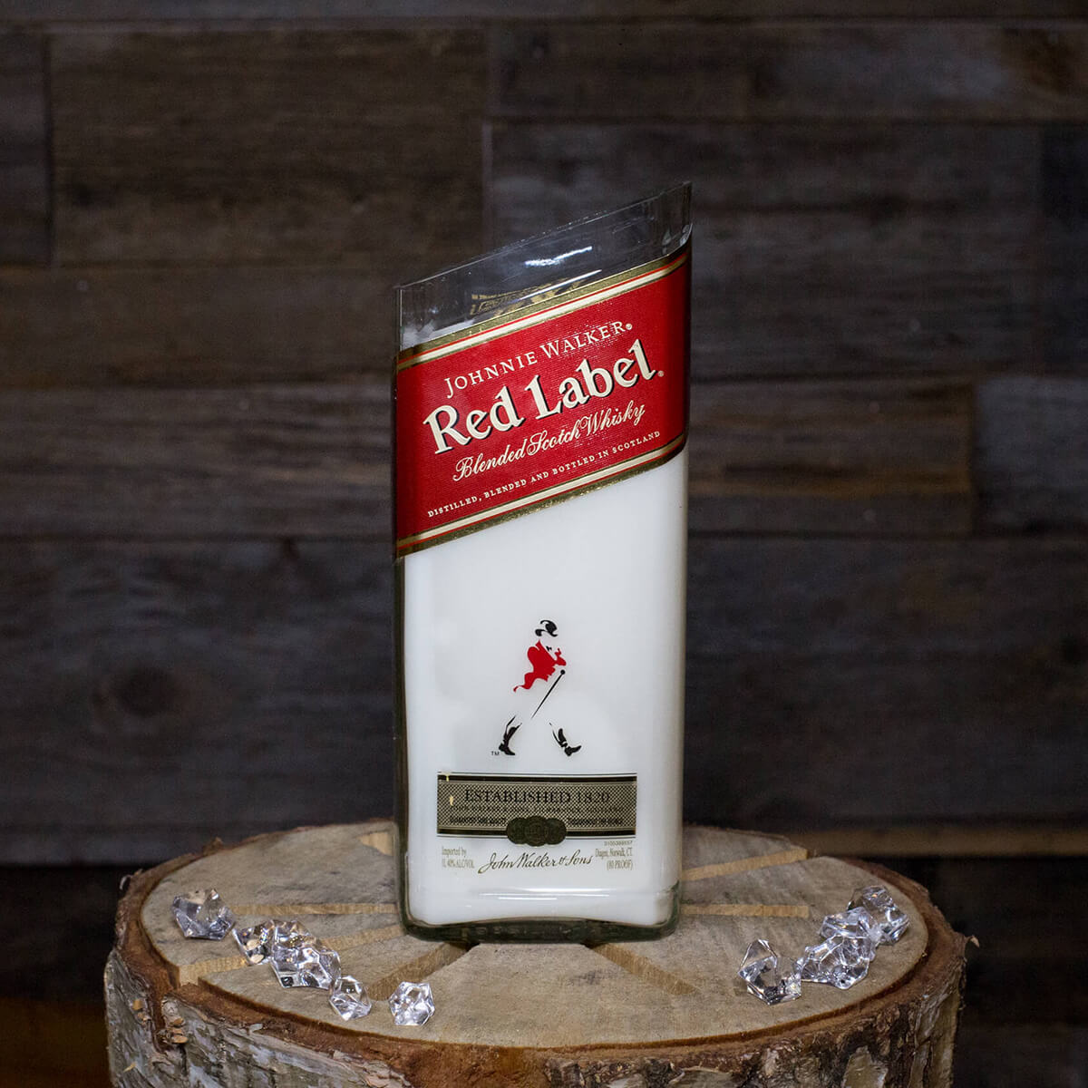 A Recycled Red Label Whiskey Candle rests on a wooden surface amid ice cubes, set against a dark wood background.