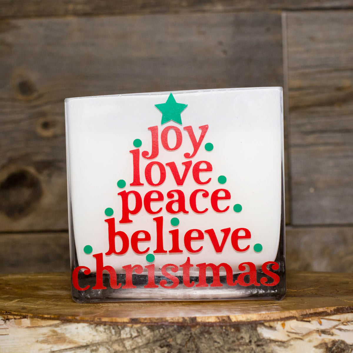 The "Frosted White Square – Love Joy Peace" candle comes in a glass holder with red and green text shaped like a Christmas tree, reading "joy love peace believe christmas," topped with a green star—ideal for festive cheer.