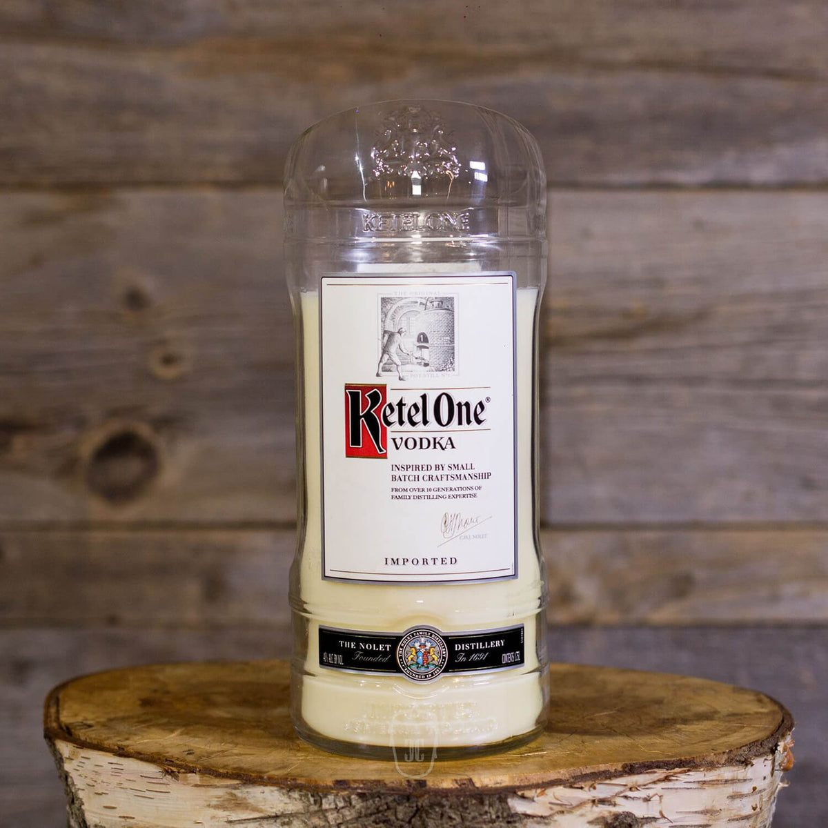 The Recycled Imported Vodka Candle, made from a repurposed vodka bottle and natural soy wax, rests on a wooden surface with a rustic wood backdrop.