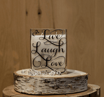 The Live Laugh Love Rectangular Candle is a white natural soy candle in a glass holder, displayed on a birch wood slice with clear decorative gems in front.