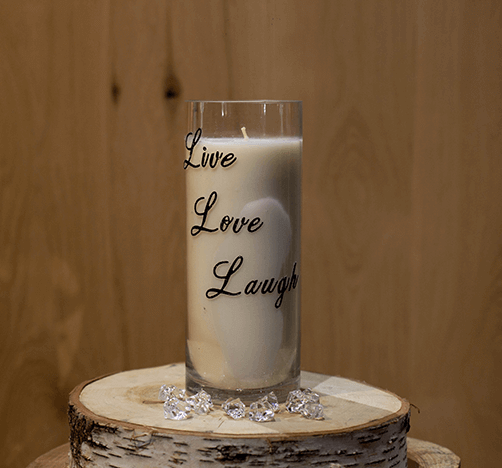The Live Love Laugh Tall Candle sits on a wooden surface, surrounded by decorative clear gems and natural soy candles, creating a cozy atmosphere.