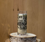 A "Love You To The Moon & Back" candle sits on a round wooden slab, surrounded by clear decorative stones and a Merry Christmas candle, filling the air with warm holiday scents.