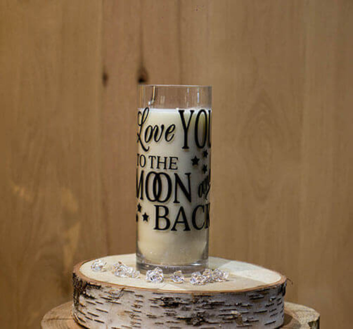 A "Love You To The Moon & Back" candle sits on a round wooden slab, surrounded by clear decorative stones and a Merry Christmas candle, filling the air with warm holiday scents.