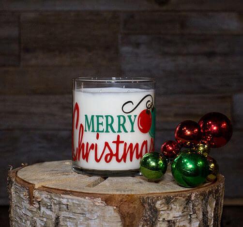 The Merry Christmas Candle, made from natural soy, sits on a cut tree stump beside red and green ornaments, set against a wooden background.