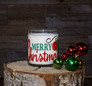 The Merry Christmas Candle, made from natural soy, sits on a cut tree stump beside red and green ornaments, set against a wooden background.