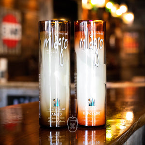 A Recycled Sunset-Colored Reposado Tequila Candle sits on a reflective wooden surface, ideal for adding unique, chic candle décor to any stylish bar or inspiring custom soy candle creations.
