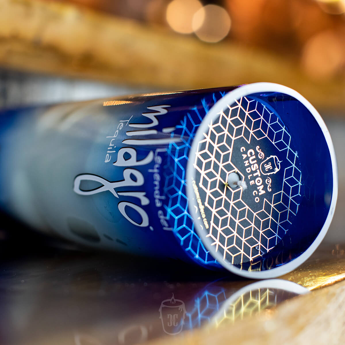 A Recycled Blue Agave Tequila Candle, crafted from a blue bottle with a geometric base, lies on its side atop a reflective surface—perfect as a stylish tequila bottle candle or agave scented home accent.