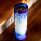 The Recycled Blue Agave Tequila Candle, in a blue and white cylindrical bottle, sits on a wooden surface near a staircase, filling the space with a rich agave scent.