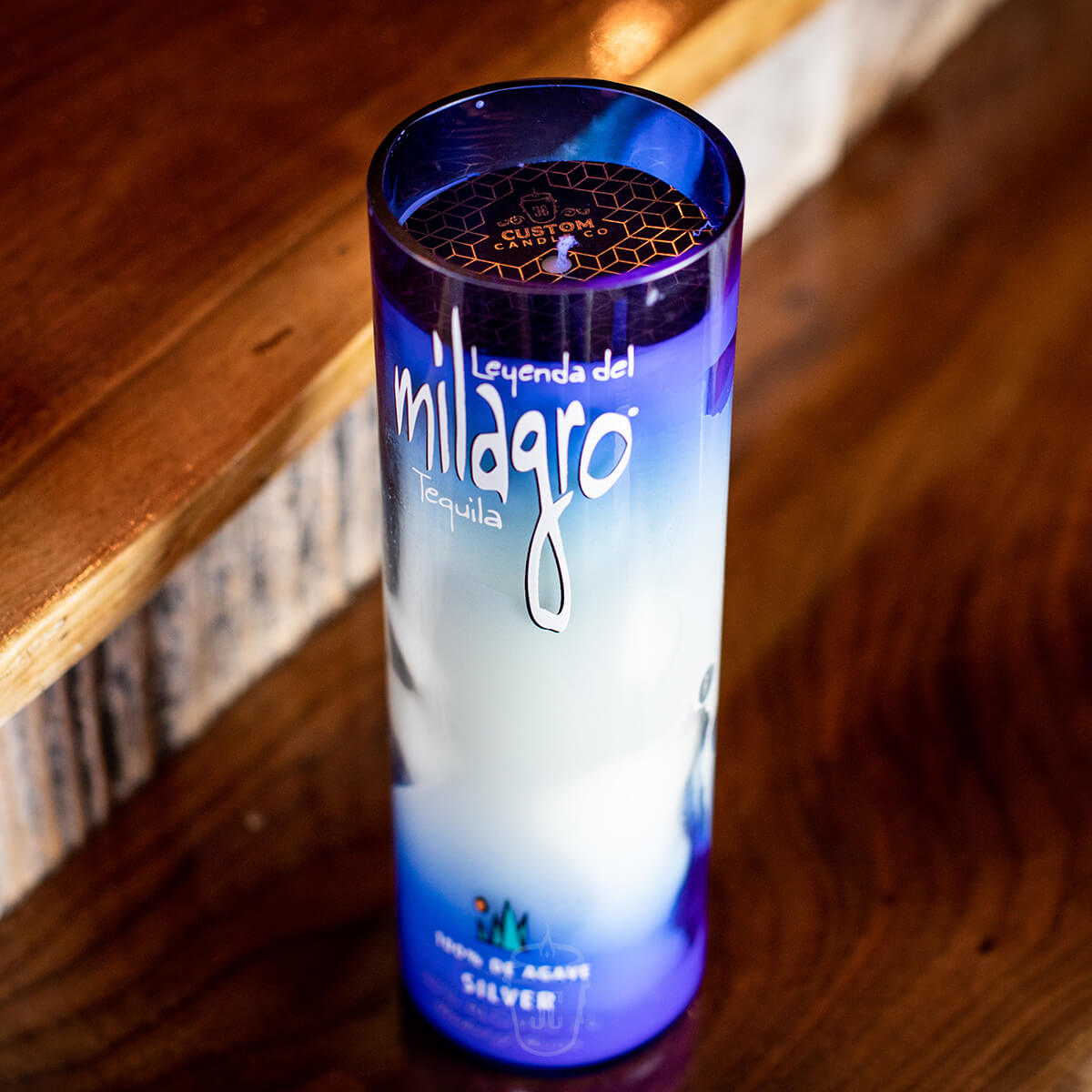 The Recycled Blue Agave Tequila Candle, in a blue and white cylindrical bottle, sits on a wooden surface near a staircase, filling the space with a rich agave scent.