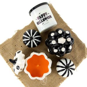 A white "Happy Halloween" soy candle, Ceramic Mini Polka Black Pumpkin Candle, black and white decorative pumpkins, and an orange candle are arranged on burlap fabric.