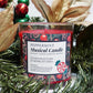 A Musical Holiday Candle – Peppermint sits among artificial greenery and gold leaves, with Christmas ornaments in the background.
