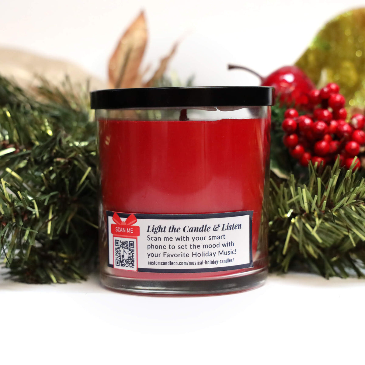 The Musical Holiday Candle & Diffuser | Peppermint features a red candle in a glass jar with a QR code on the label for holiday music, accented by artificial pine branches and red berries.
