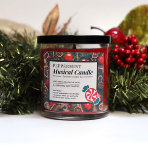 A glass jar labeled "Musical Holiday Candle & Diffuser | Peppermint" sits amid pine and red berry decor. This festive candle features cheerful graphics and a peppermint illustration—perfect for the holiday season.