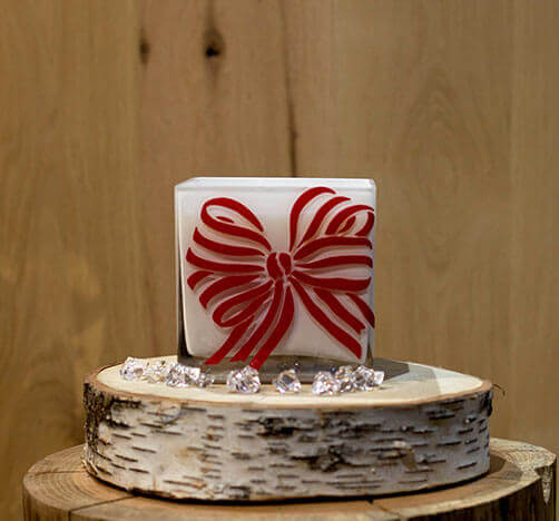 The Frosted White Square – Red Bow Candle sits on a wooden stand, surrounded by clear decorative stones and holiday-scented candles.