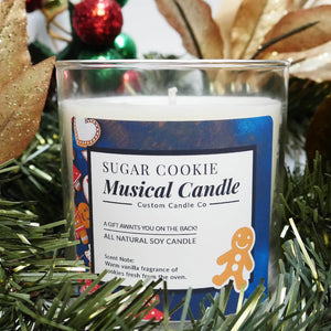 The Musical Holiday Candle – Sugar Cookie features a gingerbread man design and a warm vanilla scent, making it an ideal scented candle for the holidays, shown among green garland and festive decorations.