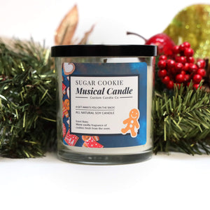 A Musical Holiday Candle & Diffuser | Sugar Cookie sits among pine branches and red berries, featuring a gingerbread man illustration on the jar.