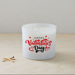 A white Happy Valentines Day Quote Candle in a frosted tumbler jar with red hearts on the glass sits on a light wooden surface beside two wooden matches.