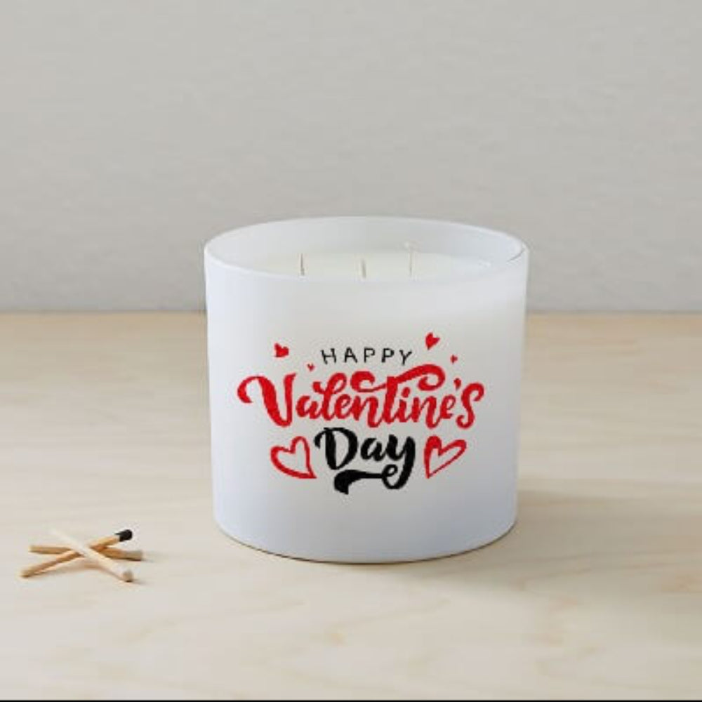 A white Happy Valentines Day Quote Candle in a frosted tumbler jar with red hearts on the glass sits on a light wooden surface beside two wooden matches.
