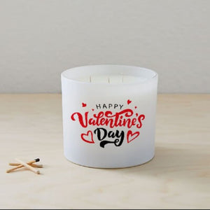 A white Happy Valentines Day Quote Candle in a frosted tumbler jar with red hearts on the glass sits on a light wooden surface beside two wooden matches.