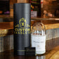 A recycled champagne candle in a clear container labeled "Whispering Angel," a branded cylindrical box, and a business card are displayed on a wooden counter.