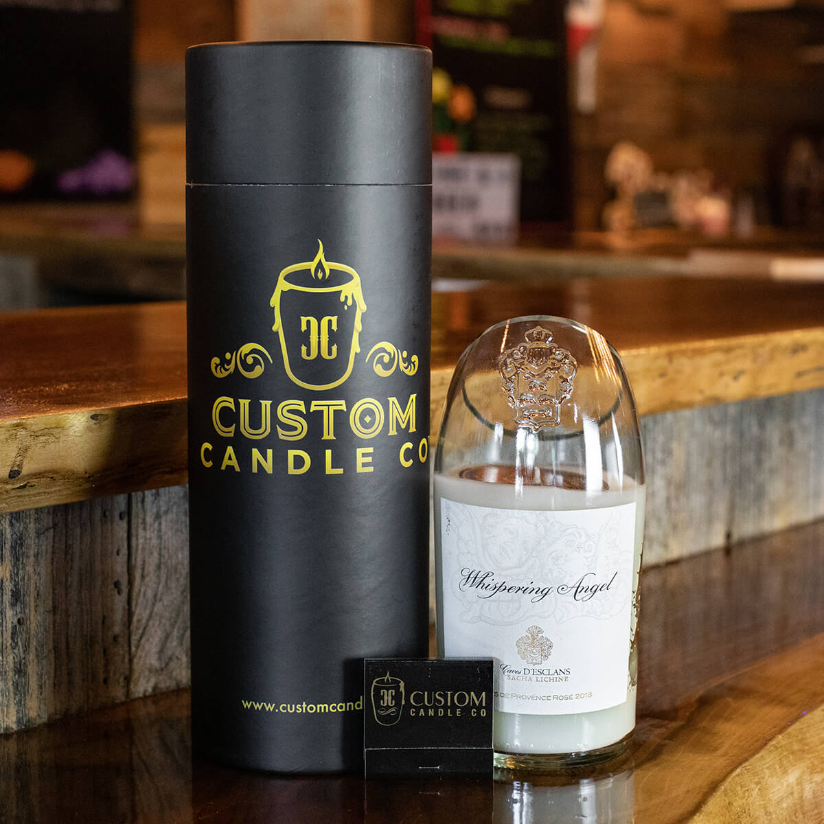 A recycled champagne candle in a clear container labeled "Whispering Angel," a branded cylindrical box, and a business card are displayed on a wooden counter.