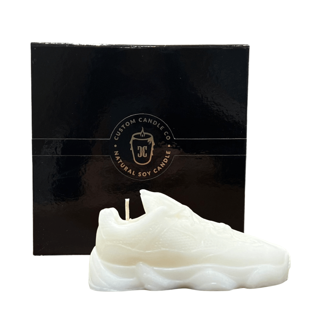 The Y500 – White Sneaker Candle – Small (On Backorder) is a hand-poured, natural soy candle shaped like a white sneaker, presented in front of a black "Custom Candle Co." box and features a refreshing Black Ice fragrance.